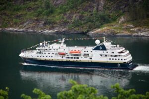 HX Expeditionsschiff MS Spitsbergen mit Flaggen geschmückt, fährt durch einen grünen Fjord mit bewaldeten Berghängen im Hintergrund.
