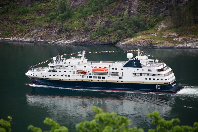 HX Expeditionsschiff MS Spitsbergen mit Flaggen geschmückt, fährt durch einen grünen Fjord mit bewaldeten Berghängen im Hintergrund.