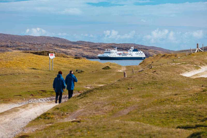 Zwei Personen wandern auf einem Pfad durch eine hügelige Küstenlandschaft, im Hintergrund liegt das HX-Expeditionsschiff MS Spitsbergen vor Anker.