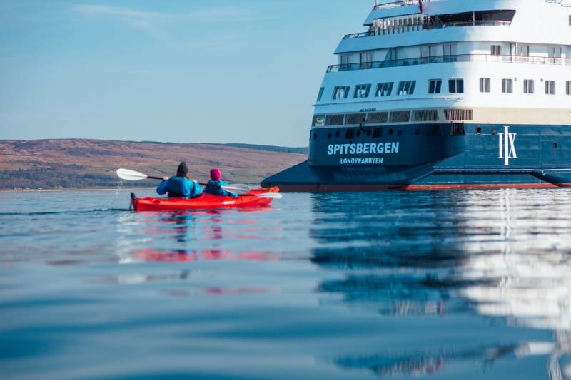 Zwei Personen paddeln in einem roten Kajak neben dem Heck des HX-Expeditionsschiffs MS Spitsbergen auf ruhigem Wasser.
