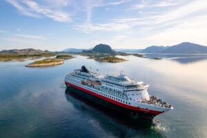 MS Finnmarken von Hurtigruten fährt durch eine malerische Insel- und Fjordlandschaft bei klarem Wetter. Spiegelglattes Wasser und Berge im Hintergrund.