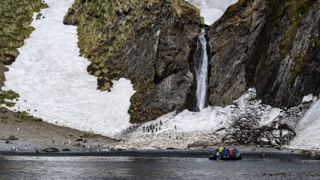 Zodiac-Boote vor einem Wasserfall in der Hercules Bay, Südgeorgien, umgeben von schneebedeckten Felsen.